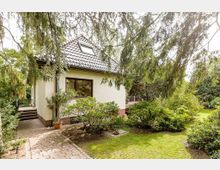 Ein kleines, weiß verputztes Einfamilienhaus mit Satteldach und einem Dachfenster, umgeben von einem üppigen Garten mit Rasen, Büschen und Bäumen. Ein gepflasterter Weg führt zum Hauseingang, und der Garten wirkt gepflegt und von Natur umgeben.