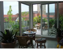 Wintergarten mit Blick auf eine begrünte Dachterrasse. Im Raum befinden sich Rattanmöbel mit Polstern, im Außenbereich Pflanzen in Töpfen, ein kleiner Tisch und Stühle; im Hintergrund sind rote Ziegeldächer und ein Kirchturm sichtbar.