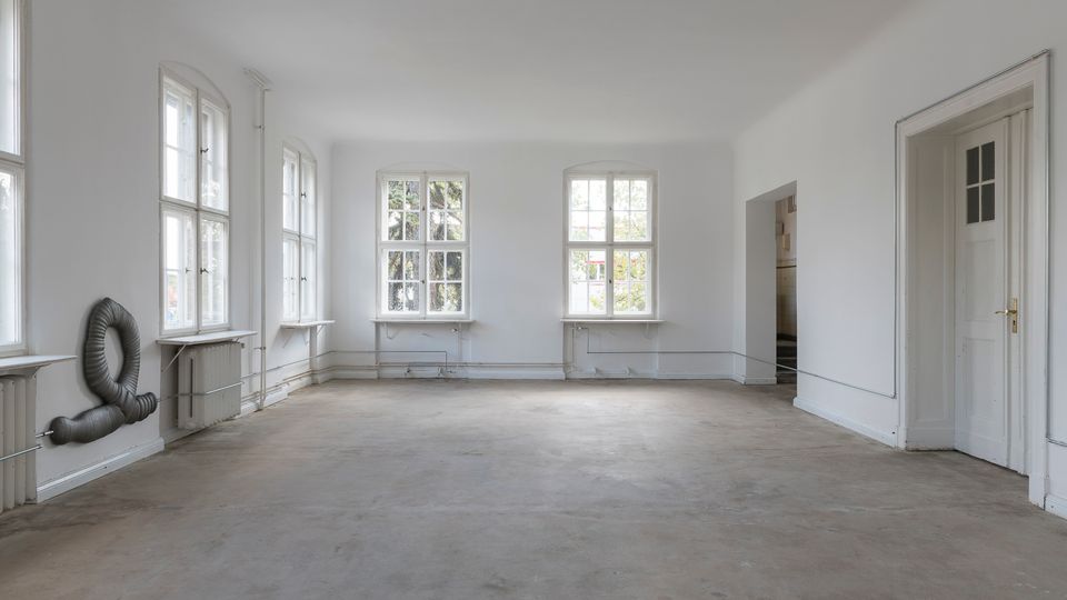 Leerstehender, heller Raum mit weißen Wänden, großen Sprossenfenstern, Betonboden und sichtbaren Heizungsrohren. Rechts befindet sich eine Tür mit goldenem Griff, und an der linken Wand ist eine graue Schlauchskulptur angebracht.