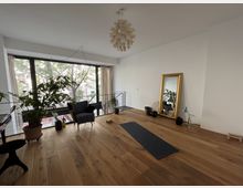 Heller Raum mit Holzboden, großen bodentiefen Fenstern und Blick auf Bäume draußen. Im Raum stehen ein Sessel, Pflanzen, ein goldgerahmter Spiegel, eine Yogamatte und Hanteln; eine moderne Hängeleuchte hängt an der Decke.