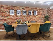 Essbereich mit einer langen Holztischplatte und bunten Samtstühlen in Gelb, Grün und Grau. Im Hintergrund eine rustikale Ziegelwand mit fünf gerahmten Schwarz-Weiß-Fotografien und einer Topfpflanze in der Ecke.