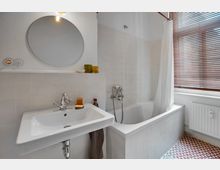 Helles Badezimmer mit einer weißen Wand, einem runden Spiegel über einem rechteckigen Waschbecken, einer Badewanne mit Duschvorhang und einer Fensterfront mit Holzjalousien. Der Boden ist mit rot-weiß gemusterten Fliesen gestaltet.