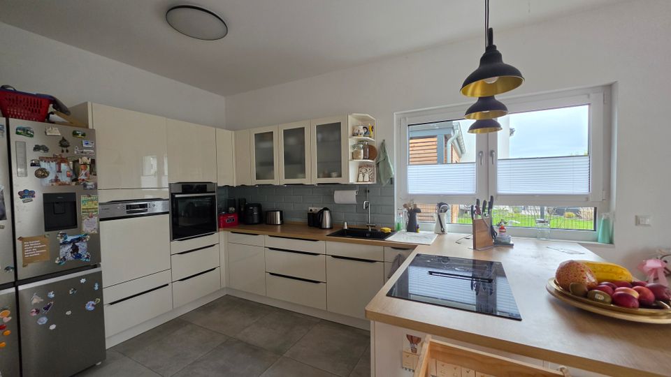 Moderne, helle Wohnküche in L-Form mit cremefarbenen Schränken, grauer Fliesenrückwand, eingebautem Backofen und großem Kühlschrank. Eine Arbeitsinsel mit Kochfeld steht vor einem breiten Fenster mit Plissees; darüber hängen schwarze Pendelleuchten.