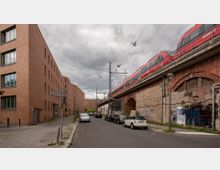 Straßenszene mit roten Regionalzügen auf einer erhöhten, gemauerten Eisenbahnbrücke rechts und modernen Backsteingebäuden links. Am Straßenrand parken mehrere Autos, und vereinzelte Graffiti befinden sich an den Brückenwänden.