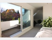 Schlafzimmer mit großen Schiebetüren aus Glas, die zu einem Balkon mit Holzboden und Pflanzen führen. Im Hintergrund sind Bäume mit Herbstlaub und ein Haus mit rotem Ziegeldach sichtbar.