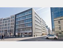 Moderne Bürogebäude an einer Straßenecke mit Glasfassaden und rechteckigen Fenstern, umgeben von weiteren urbanen Gebäuden. Im Vordergrund sind Gehwege, Autos und mehrere Passanten zu sehen.