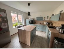 Moderne Küche mit mintfarbenen Schränken und Holzarbeitsflächen. In der Mitte steht eine Kücheninsel, links ein Kühlschrank, rechts eine Spüle, und darüber hängt eine schwarze Deckenleuchte.