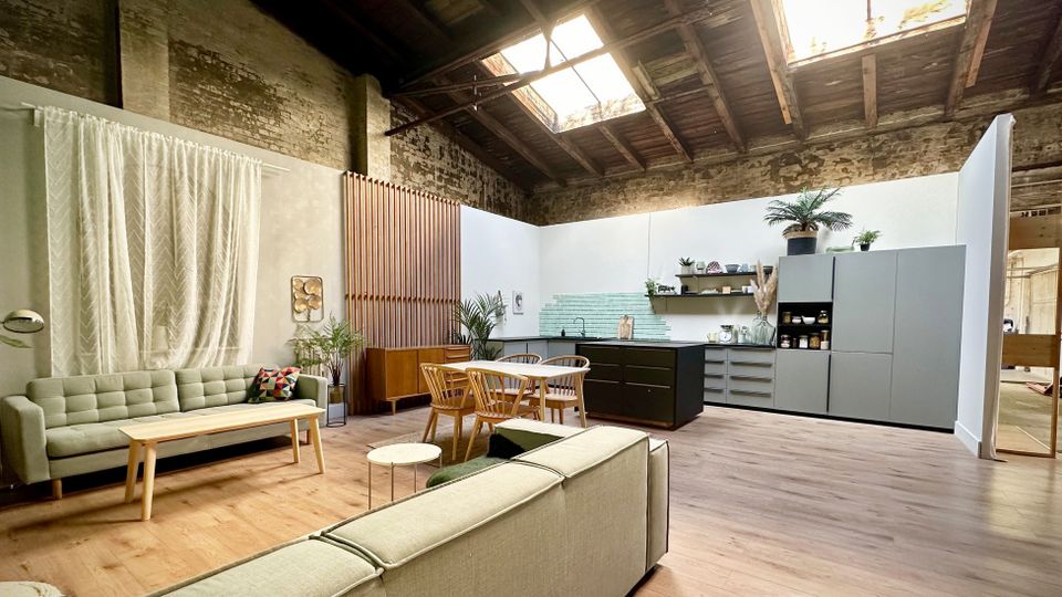 Offener Loftraum mit hoher Decke, sichtbaren Holzbalken und großen Oberlichtern sowie freigelegten Backsteinwänden. Im Vordergrund ein Wohnbereich mit Sofas und Couchtisch, daneben ein Esstisch und eine moderne Küchenzeile mit Regalen und Pflanzen.