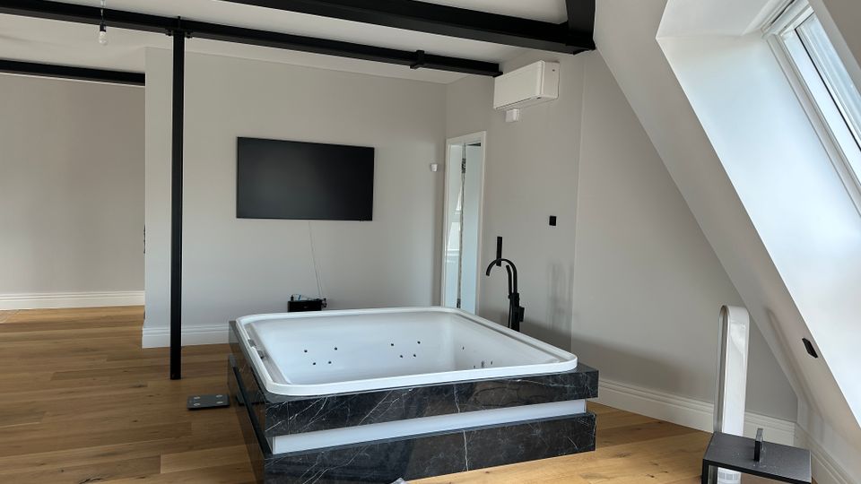 Modernes Dachgeschosszimmer mit freistehender Whirlpool-Badewanne auf dunklem Marmorsockel, Holzfußboden und sichtbaren schwarzen Deckenbalken. Rechts sorgt ein großes Dachfenster in der Schräge für Tageslicht; an der Wand hängen ein Fernseher und eine Klimaanlage.