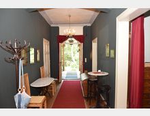 Ein schmaler Flur mit dunklen Wänden, auf dem ein roter Teppich liegt. Links stehen Holzmöbel mit kleinen Marmortischen, rechts ein runder Stehtisch und Barhocker; am Ende des Flurs führt eine Tür mit roten Vorhängen ins Freie, mit Blick auf eine grüne Außenanlage.