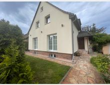 Zweistöckiges Einfamilienhaus mit cremefarbener Fassade, dunklem Dach und Ziegelverkleidung im unteren Bereich. Vor dem Haus befindet sich ein gepflegter Garten mit Rasen, Sträuchern und einem gepflasterten Weg, der zum Eingang führt.