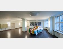 Modernes Foyer mit gefliesten Böden und weißen Wänden, beleuchtet von Wandlampen. Im Raum steht ein bunt bemalter Oldtimer, und an einer Wand hängen schwarz-weiße Fotografien; große Fenster lassen Tageslicht hinein.