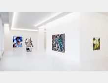 Innenansicht einer modernen Kunstgalerie mit weißen Wänden und hellem Boden. An den Wänden hängen drei Gemälde in verschiedenen Stilen, und in der Mitte des Raums steht eine skulpturale Installation.