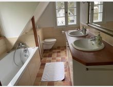 Badezimmer unter einer Dachschräge mit Badewanne links, wandhängendem WC vor einem Fenster und Waschtisch mit zwei Waschbecken rechts. Beige Wandfliesen, rot-braune Bodenfliesen und ein großer Spiegel über dem Waschtisch prägen den Raum.
