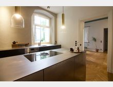 Moderne Küche mit einer zentralen Kochinsel aus Edelstahl, einem integrierten Induktionskochfeld und hängenden Glasleuchten. Im Hintergrund ein großes Fenster mit Rundbogen, dunkle Einbauschränke, und ein Durchgang mit Blick auf einen Raum mit Parkettboden und einer Pflanze.