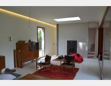 Modernes Wohnzimmer mit minimalistischem Design. Der Raum hat weiße Wände, einen Kamin mit schwarzer Verkleidung, ein Oberlicht, Holzstühle mit Polstern, ein roter Teppich sowie eine Vitrine aus Holz. Ein großes Fenster bringt Tageslicht herein, und im Hintergrund ist ein angrenzender Raum sichtbar.
