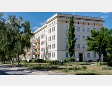 Ein mehrstöckiges, helles Wohngebäude mit verzierten Fassadenelementen und Balkonen, umgeben von Bäumen und einer Grünfläche. Im Vordergrund verläuft ein gepflasterter Weg, und eine Person mit Fahrrad ist zu sehen.