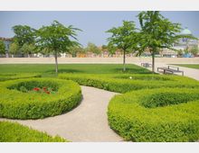 Ein gepflegter Park mit grünen Hecken in geschwungenen Formen, vereinzelt stehenden Bäumen und roten Blumen im Vordergrund. Im Hintergrund sind ein gepflasterter Platz, Bankreihen, und historische Gebäude mit einer Kuppel zu erkennen.