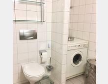 Gefliestes Badezimmer mit wandhängender Toilette, Edelstahl-Spültaste und Glasablage an der Wand. In einer Nische steht eine Frontlader-Waschmaschine neben einem Metall-Wäschebehälter auf hellen Bodenfliesen.