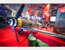 Ein Ausstellungsraum mit einem historischen Jagdflugzeug auf einem roten Teppich, umgeben von grünen Fässern und Absperrungen. Im Hintergrund befinden sich Werbebanner mit der Aufschrift „War Thunder“ sowie zahlreiche Besucher an verschiedenen Ständen.