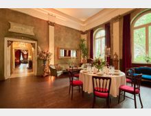 Eleganter Raum mit klassisch dekoriertem Essbereich, bestehend aus einem runden Tisch mit rotsamtbezogenen Stühlen und weißer Tischdecke, geschmückt mit Kerzenleuchtern und Blumenarrangements. Hohe Fenster mit roten Vorhängen, eine antike Uhr und eine Sitzecke mit Möbeln im Vintage-Stil prägen das Ambiente, während im Hintergrund ein opulenter Kronleuchter durch eine offene Tür sichtbar ist.