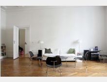 Helles Wohnzimmer mit weiß gestrichenen Wänden, einem Parkettboden, einem weißen Sofa mit grünen Kissen, mehreren schwarzen Stühlen und einem runden Couchtisch. Im Hintergrund steht ein offenes Bücherregal und eine geöffnete Tür führt zu einem weiteren Raum.