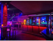 Innenraum einer Bar oder eines Clubs mit neonfarbener Beleuchtung in Blau, Rot und Violett, langer Theke mit beleuchteter Front und Regalen mit Flaschen. Sichtbare Ziegelpfeiler, hohe Decke mit Lüftungsrohr sowie Sitznischen, Stehtische und Barhocker im Loungebereich.