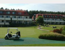 Eine Hotelanlage mit roten Dächern und mehreren Etagen, umgeben von einem gepflegten Rasen und einer Golfanlage. Im Vordergrund steht ein Golfcart, während im Hintergrund Personen mit Golf-Ausrüstung auf einem Weg unterwegs sind, daneben grast eine Kuh.