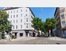 Straßenkreuzung in einem Wohngebiet mit mehrstöckigen Altbauhäusern, teilweise renoviert. Die Straße ist von Bäumen gesäumt, mit gepflastertem Boden und Verkehrsschildern, darunter Einfahrt-Verboten-Schilder.