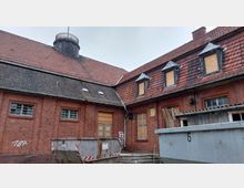 Backsteinbau mit roten Dachziegeln und mehreren Gaubenfenstern, die größtenteils mit Sperrholzplatten abgedeckt sind. Im Vordergrund befinden sich ein Metallcontainer sowie eine Treppe, die zu einer verschlossenen Tür führt.