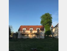 Zweistöckiges Wohnhaus mit weißer Fassade und rotem Ziegeldach, ausgestattet mit einem hölzernen Balkon vor mehreren Fenstern im Obergeschoss, umgeben von einer Wiese und einem großen Baum. Rechts im Hintergrund ist ein weiteres Gebäude mit einer kleinen Turmspitze sichtbar.