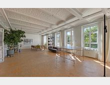 Heller, offener Raum mit weiß gestrichenen Backsteinwänden und sichtbaren Deckenbalken, großen Fenstern, die Tageslicht hereinlassen, sowie Holzboden. Der Raum ist möbliert mit Tischen, Stühlen, einem Regal, einem Sofa und einer großen Topfpflanze.