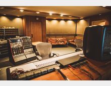 Aufnahme eines Tonstudios mit einem großen Mischpult im Vordergrund, Regale mit technischen Geräten an der Wand und braunen Ledersofas im Hintergrund. Die Wände sind mit Holzvertäfelung und schallabsorbierenden Paneelen verkleidet, und eine warme Deckenbeleuchtung erzeugt eine gleichmäßige Ausleuchtung des Raumes.