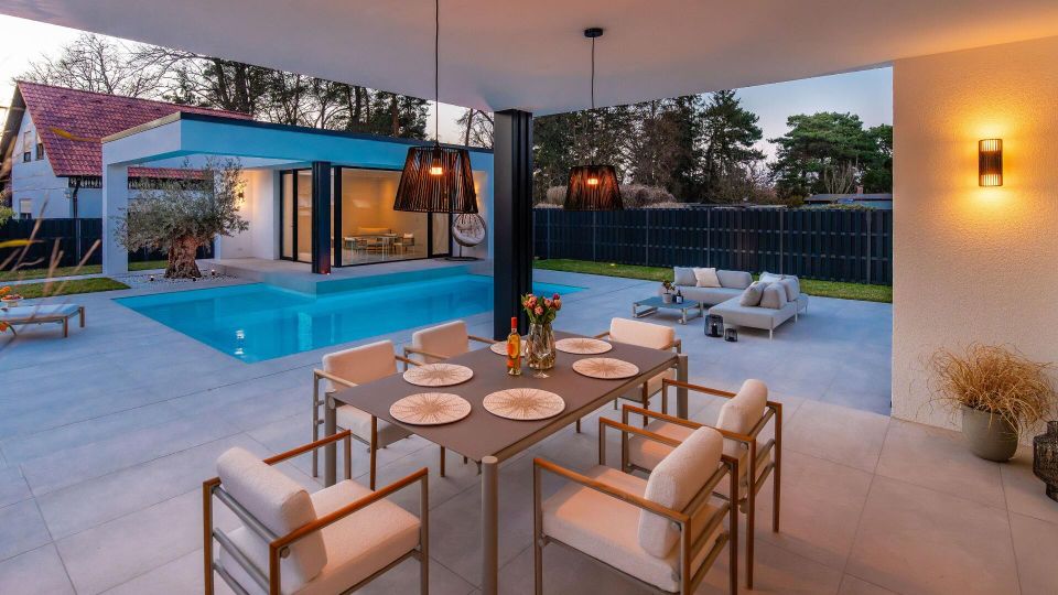 Überdachte Terrasse eines modernen Wohnhauses mit großformatigen Fliesen, Esstisch und Loungemöbeln, mit Blick auf einen rechteckigen Pool. Im Hintergrund sind bodentiefe Glasfronten, eine überdachte Sitznische, dunkler Sichtschutzzaun und Gartenbepflanzung zu sehen.