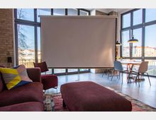 Wohnraum mit moderner Einrichtung, bestehend aus einer roten Couch, passenden Sesseln und einem Tisch mit Stühlen im Hintergrund. Große Fenster lassen viel Tageslicht herein, und eine heruntergelassene Leinwand befindet sich zentral vor der Glasfront.