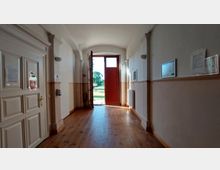 Heller Flur mit Holzboden, hohen Wänden und einem großen, offenen Holztürrahmen, der nach draußen führt. Durch die geöffnete rote Tür ist ein sonniger Garten mit Bäumen sichtbar.