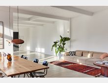 Offener Wohnraum mit minimalistischem Design, bestehend aus einer hellen Polstercouch, einem Holztisch mit schwarzen Stühlen, einer großen Zimmerpflanze und einem roten Teppich. Weiße Wände, sichtbare Deckenbalken und ein modernes Pendellicht ergänzen das Interieur, das von natürlichem Licht durchflutet wird.