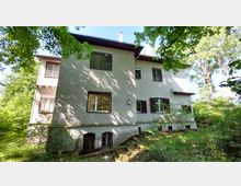 Zweistöckiges Wohnhaus mit verputzter Fassade, großen Fenstern und teils geschlossenen Fensterläden, umgeben von dichtem Grün und Bäumen. Im Vordergrund befindet sich ein kleiner, leicht überwucherter Garten mit einem niedrigen Zaun.