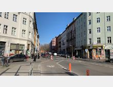 Straßenszene in einer urbanen Umgebung mit mehrstöckigen Wohn- und Geschäftshäusern, teils mit Graffiti bedeckt. Im Vordergrund ist eine Kreuzung mit Fahrradwegmarkierungen und parkenden Autos sowie Passanten auf den Gehwegen zu sehen.