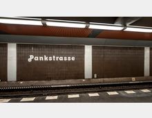 U-Bahn-Station mit der Aufschrift „Pankstraße“ an einer Wand aus dunkelbraunen Fliesen. Die Gleise und der Bahnsteig mit einem Zebrastreifenmuster im Haltebereich sind sichtbar, die Decke ist mit orangefarbenen Paneelen gestaltet.