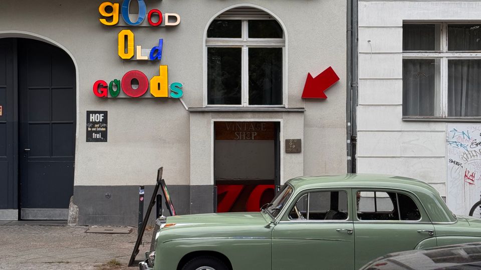 Fassade eines Altbaugebäudes mit einem Ladenlokal mit der Aufschrift „Möbel“ in grüner Leuchtschrift und einem bunten „Good Old Goods“-Schriftzug. Vor dem Gebäude sind ein grüner Oldtimer und ein schwarzes Auto auf der Straße geparkt.