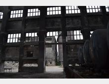 Innenansicht einer verlassenen Industriehalle mit hohen Zementwänden und großen Fensteröffnungen ohne Glas. Im Hintergrund sind eine Ruine und ein alter Schornstein zu sehen, während im Vordergrund rostige Tanks und Trümmer liegen.