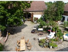 Ein gepflasterter Innenhof mit rustikalem Charme, umgeben von Bäumen und Pflanzen. Es sind Holzstühle und ein langer Tisch, eine Feuerstelle, sowie Blumenbeete, eine Schaukel und Sitzgelegenheiten sichtbar; im Hintergrund steht ein Gebäude mit roten Ziegeln und großen Toren.