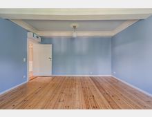 Leerer Wohnraum mit hellblauen Wänden, breiten weißen Stuckleisten an der Decke und einer zentralen Deckenlampe. Holzdielenboden und offene weiße Tür zu einem angrenzenden Flur, mit Klimagerät über der Tür.