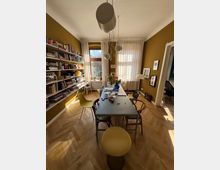 Esszimmer mit Holzboden, gelb gestrichenen Wänden und einem langen Tisch, umgeben von verschiedenen Stühlen. Links befinden sich Bücherregale an der Wand, vor einem großen Fenster mit weißen Vorhängen stehen Pflanzen und Dekorationen. Eine moderne Lampe hängt von der Decke.