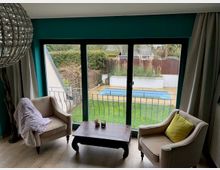 Wohnzimmer mit zwei gepolsterten Sesseln und niedrigem Couchtisch vor einer großen, dreiteiligen Glas-Schiebetür. Durch die Fensterfront sieht man einen kleinen Garten mit Rasen, gepflasterter Terrasse und einem schmalen Pool, eingefasst von Zaun und Hecken.