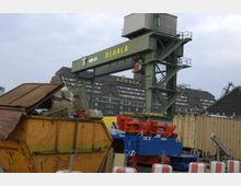 Ein Industriehafen mit einem grünen Verladekran der Firma "Behala". Im Vordergrund stehen ein gelber Container mit Schrott und blaue sowie rote Metallteile; im Hintergrund sind Backsteingebäude mit der Aufschrift "Behala" sichtbar.