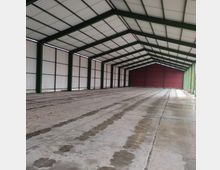 Große, leere Lagerhalle mit einem Dach aus weißem Wellblech und grünen Stahlträgern. Die Wände und das Tor am Ende der Halle bestehen aus Metallplatten, der Boden aus Beton mit Wasserflecken.