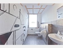 Kleines Badezimmer mit grauen Fliesen und einem Wand-WC. Links zeigt eine künstlerisch bemalte Wand abstrakte Schwarz-Weiß-Motive, während das rechteckige Fenster am Ende des Raumes Tageslicht hineinlässt.