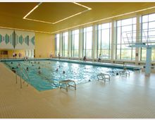 Hallenbad mit großem Schwimmbecken, umgeben von beige Fliesen. Große Fensterfronten lassen Tageslicht herein, und an der Wand ist ein grafisches Mosaikmuster zu sehen; im Wasser schwimmen Menschen, während andere am Rand stehen oder sitzen.